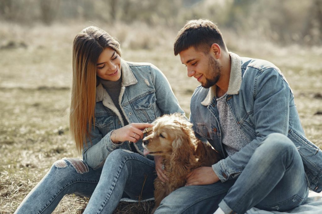 Happy young couple sitting on plaid with dog on sunny field in countryside